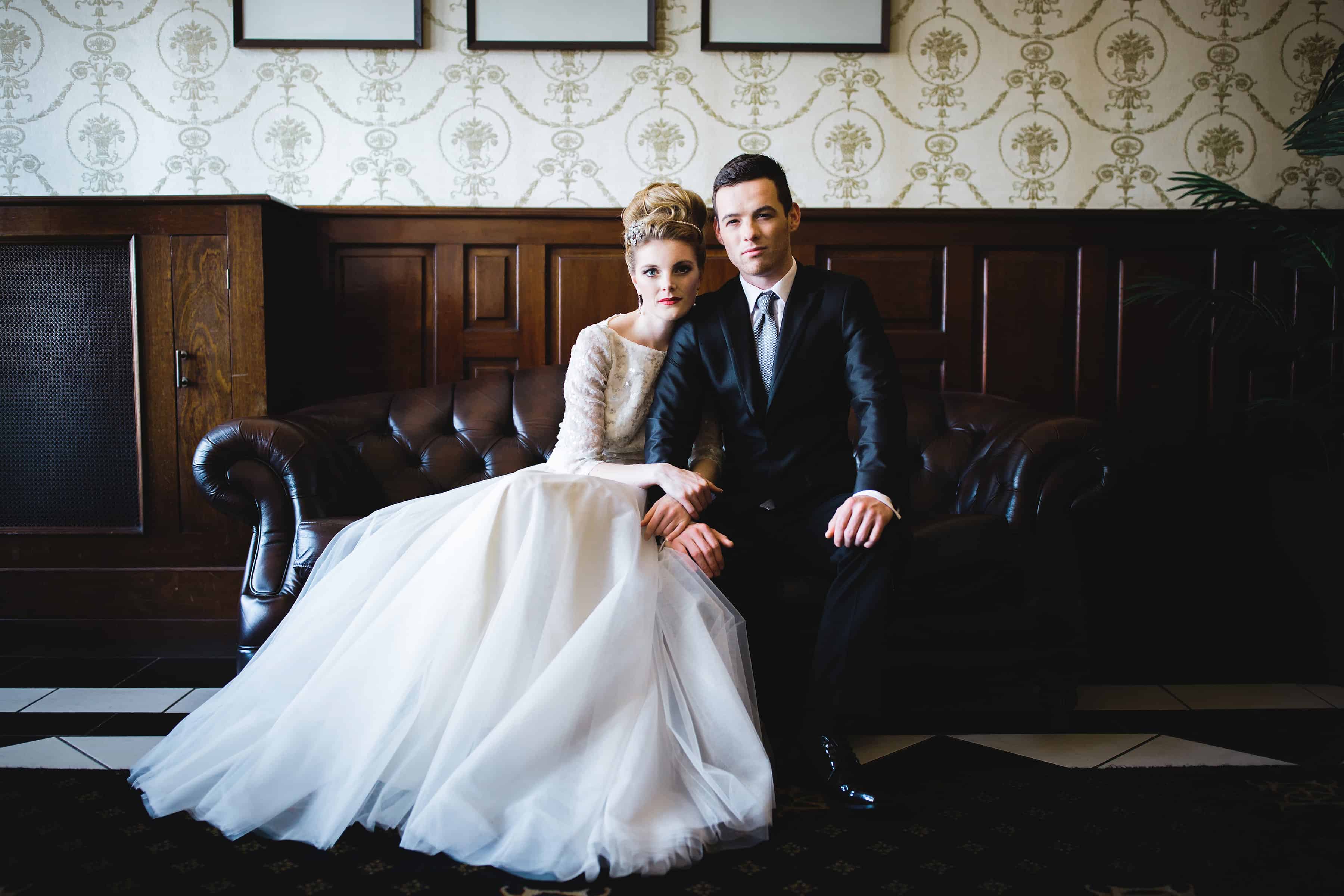 A bride and groom sitting on a sofa at the Duke of Cornwall Hotel, with the bride wearing wedding jewellery