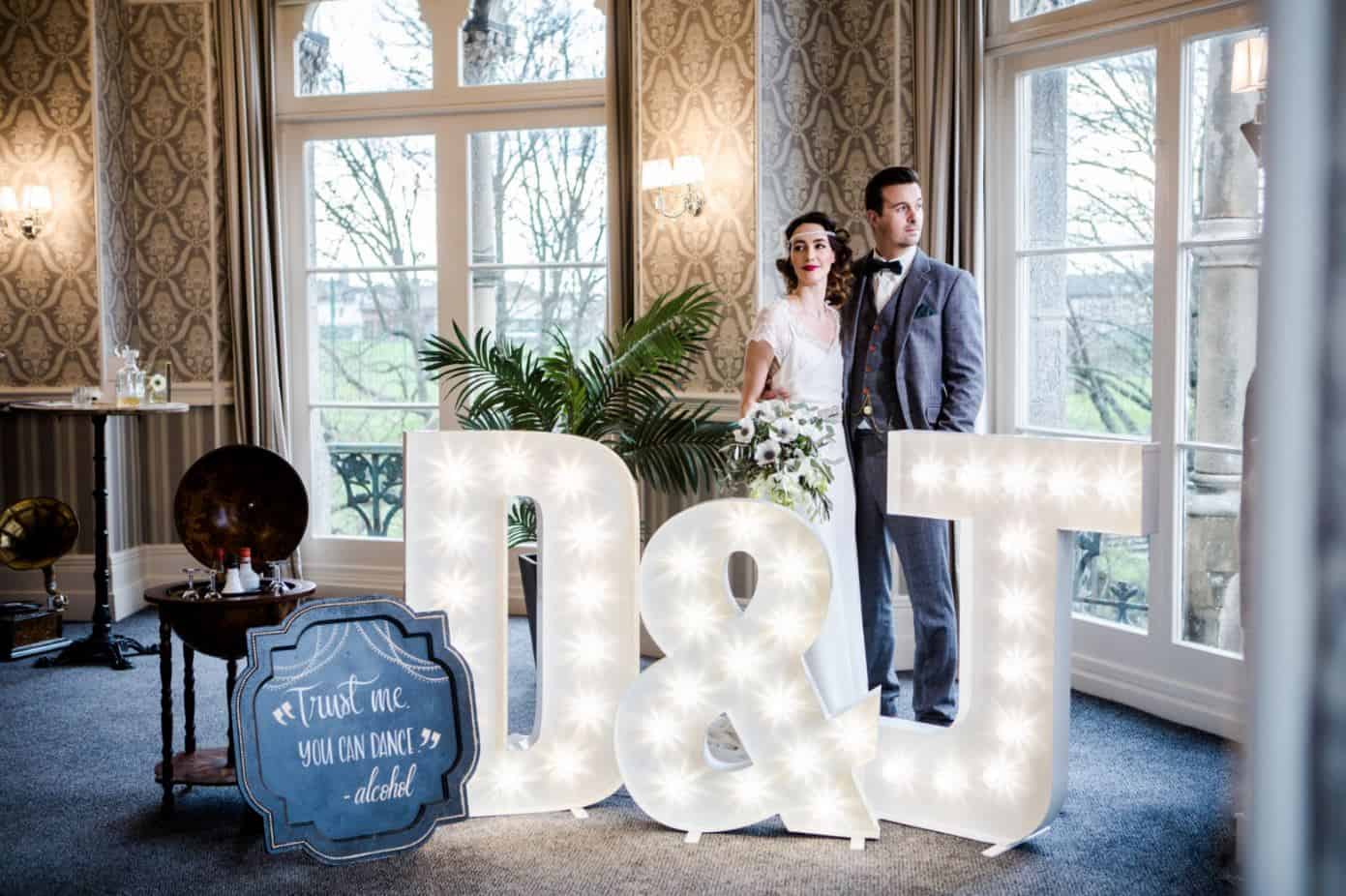 Bride and groom standing with wedding props, including lighted letters and a vintage sign